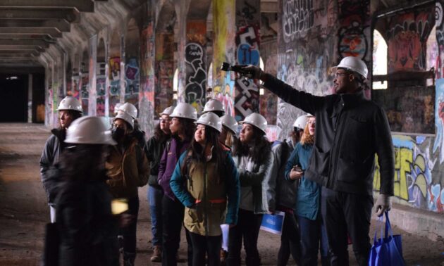 Man in hard hat directs a group of students in hard hats touring the City of Rochester's graffiti-lined abandoned aqueduct underground subway.