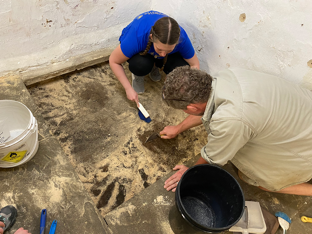 A student and a professor kneel over a square hole in a structure, scraping the surface with archeological tools.