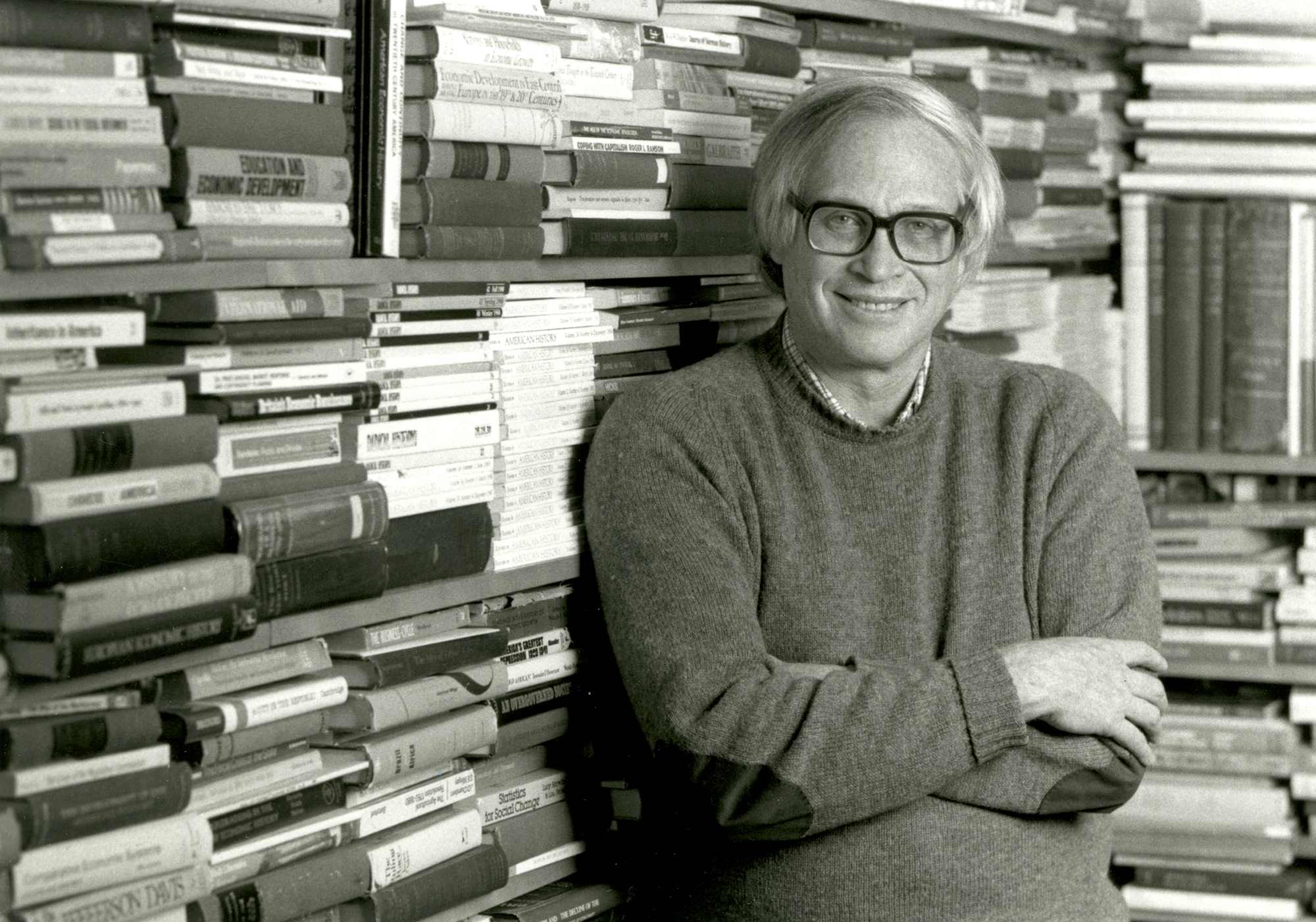 archival photo of Stanley Engerman, standing in front of a large bookshelf.