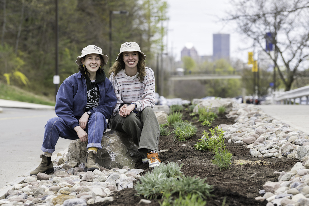 Two women students siting on large rock surrounded by fresh mulch and new plantings.