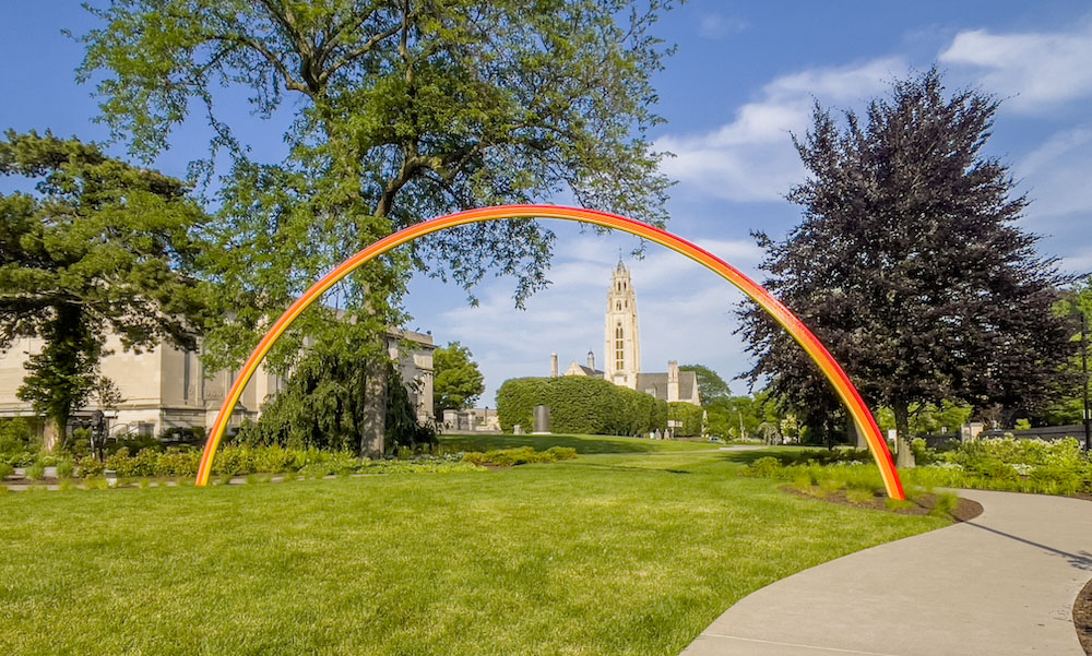 Rainbow sculpture framing MAG tower against blue sky.