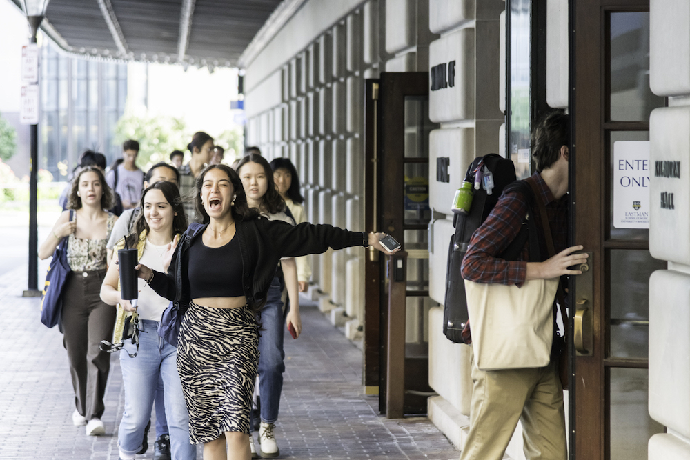 A number of students heading into a school building. Student in front is smiling, arms open. 