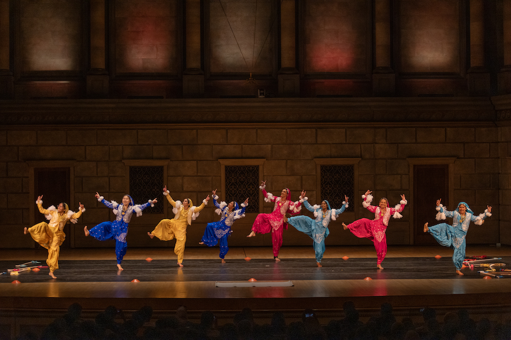 Eight members of the Bhangra dance team on a stage in royal blue, gold, pink, and turquoise outfits. 