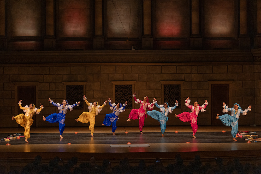 Eight members of the Bhangra dance team on a stage in royal blue, gold, pink, and turquoise outfits. 