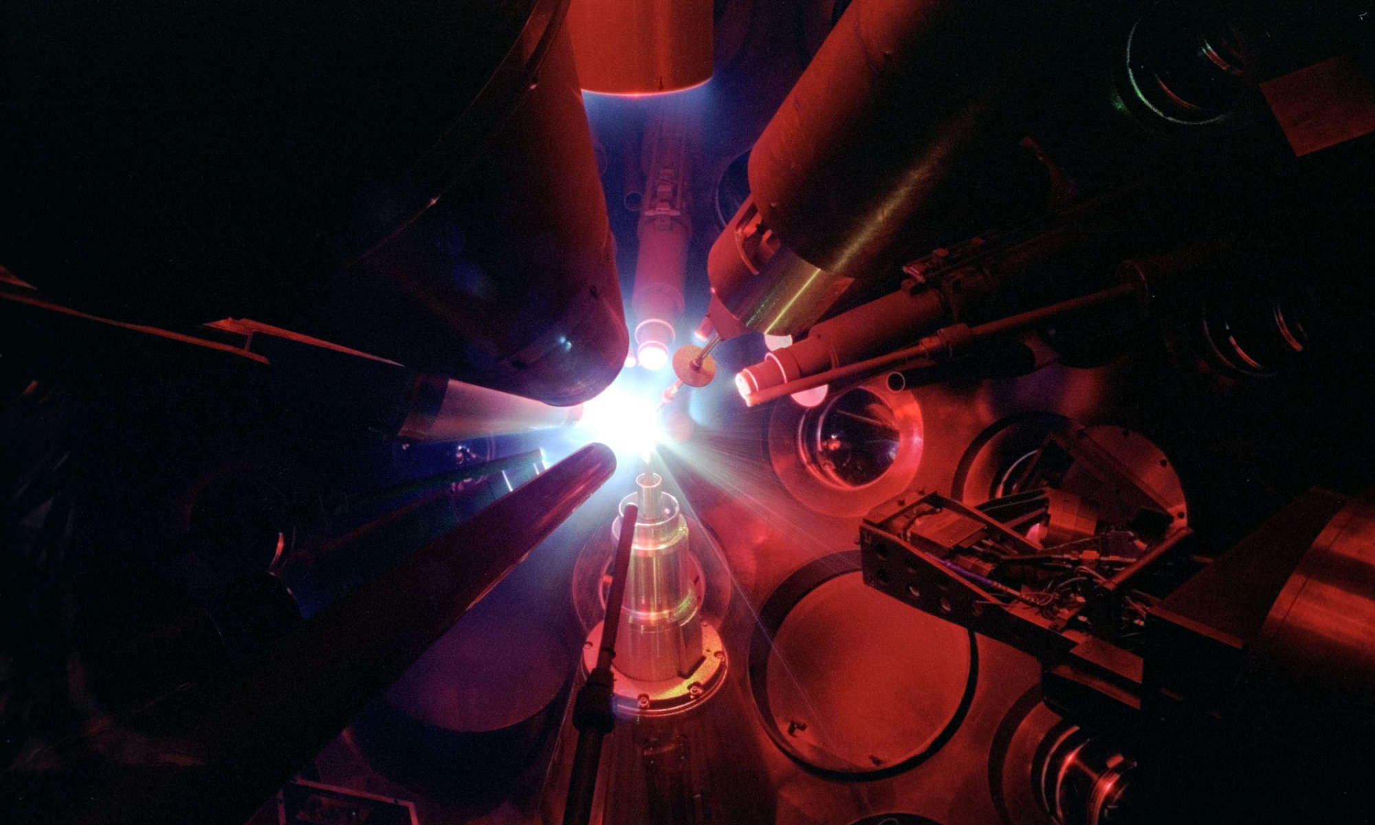 Inside the OMEGA target chamber, multiple lasers are aimed at a target amid a red cast of light.