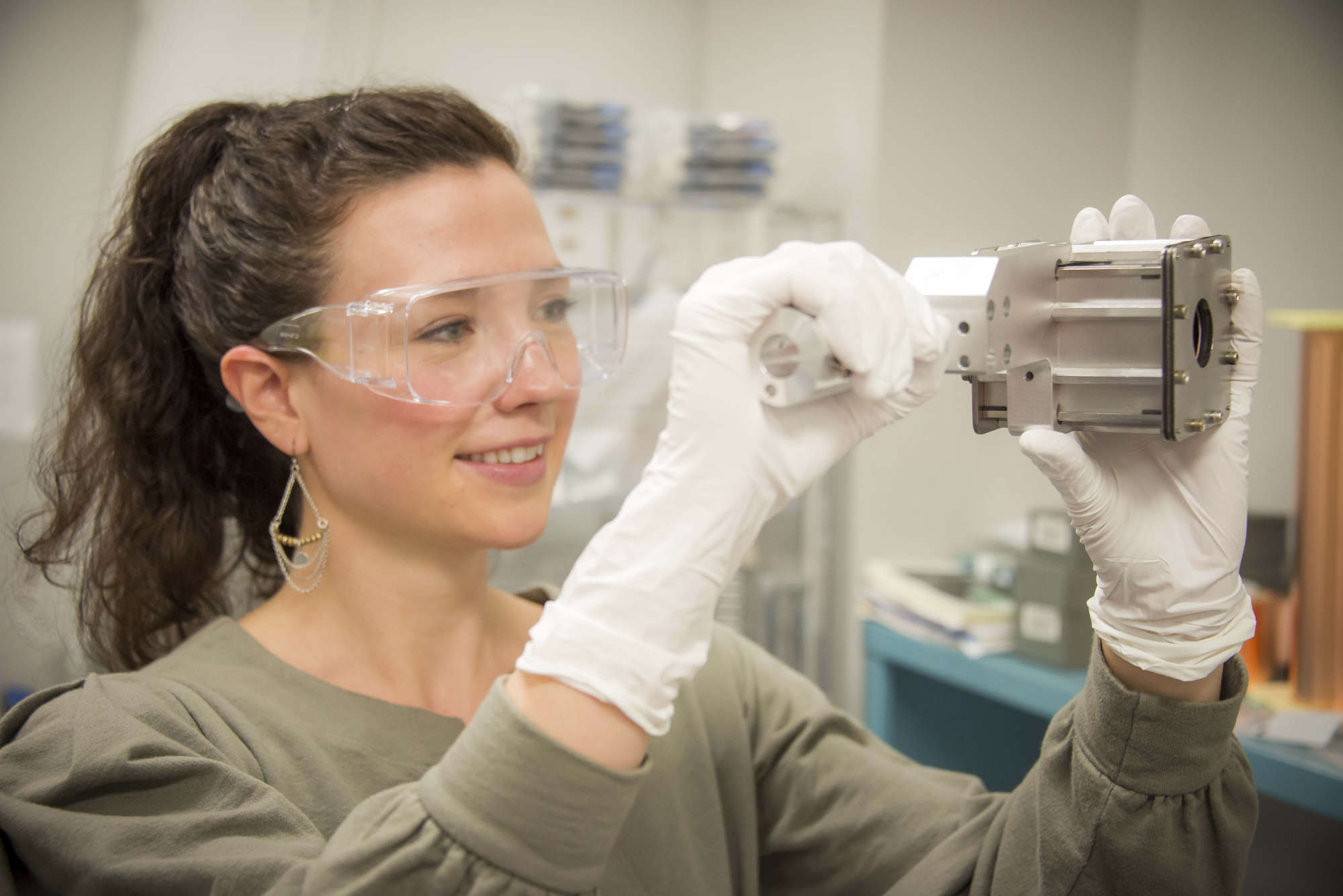 Danae Polsin in safety goggles and gloves holds up and analyzes a piece of lab technology.