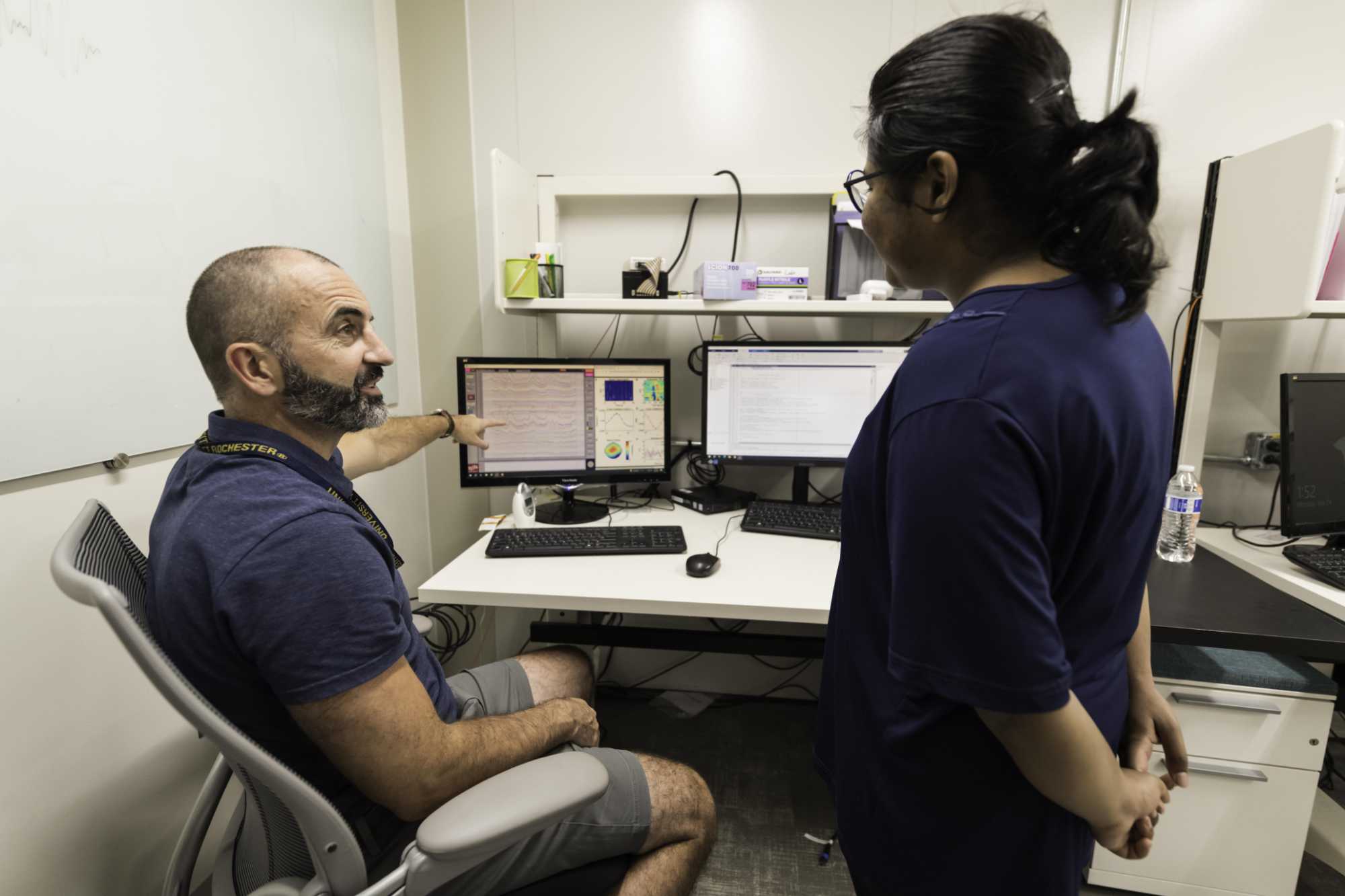 An undergraduate student stands and speaks to a seated professor in a neuroscience lab with computer screens displaying research data.