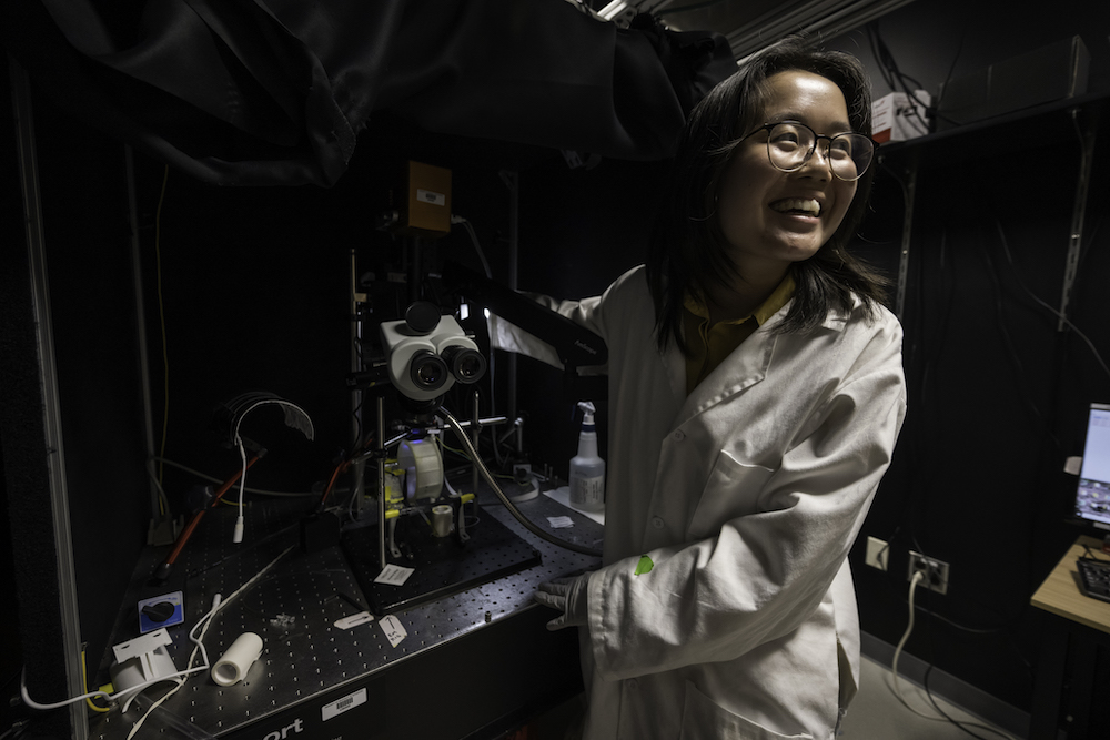 Student in white lab coat smiles, surrounded by lab equipment