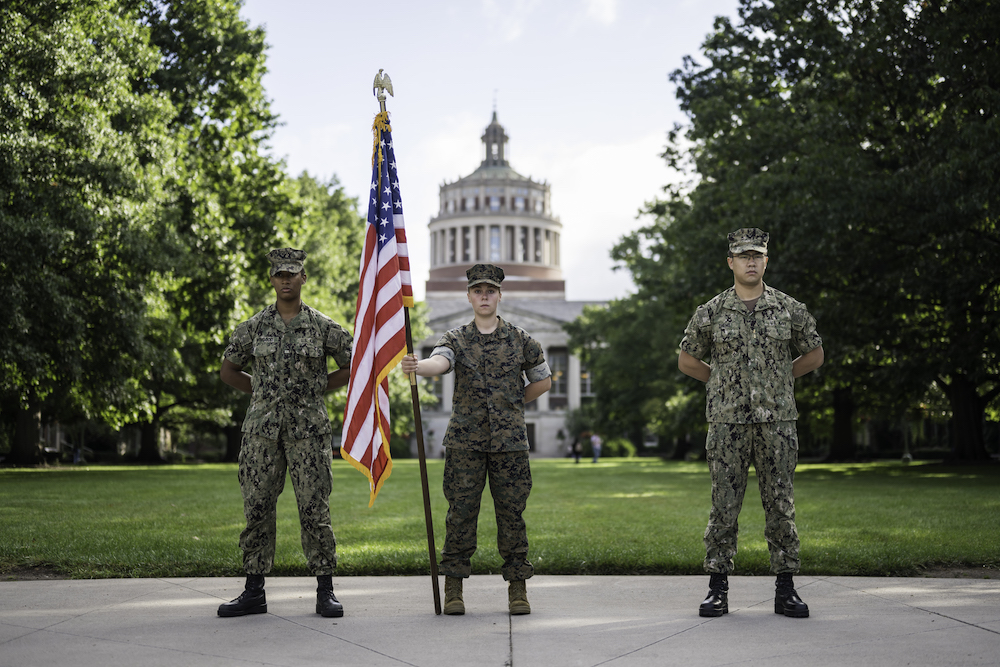 Three people in military fatigues stand at attention with an American flag