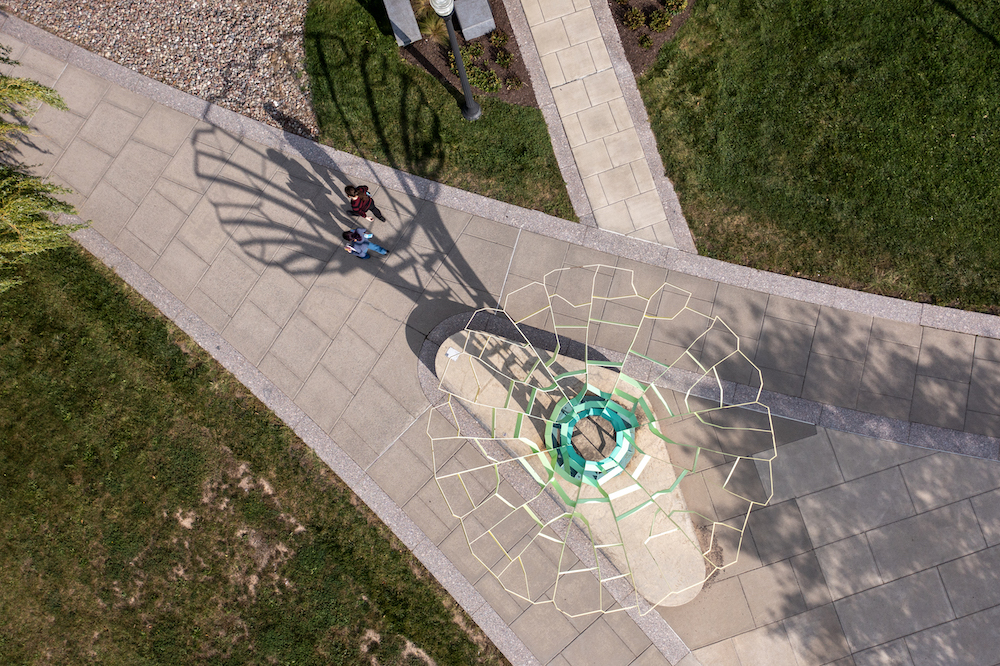 Overhead view of students walking along a paved path, grass on either side. A shadow from a sculpture is cast over them.