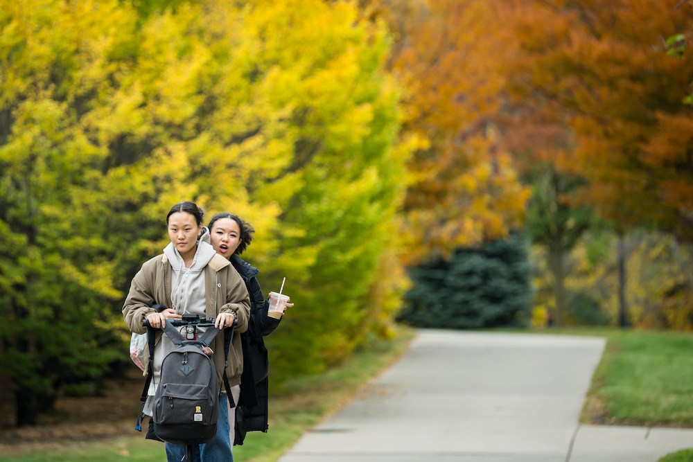 Two students, one with coffee in hand, ride a scooter through fall foliage