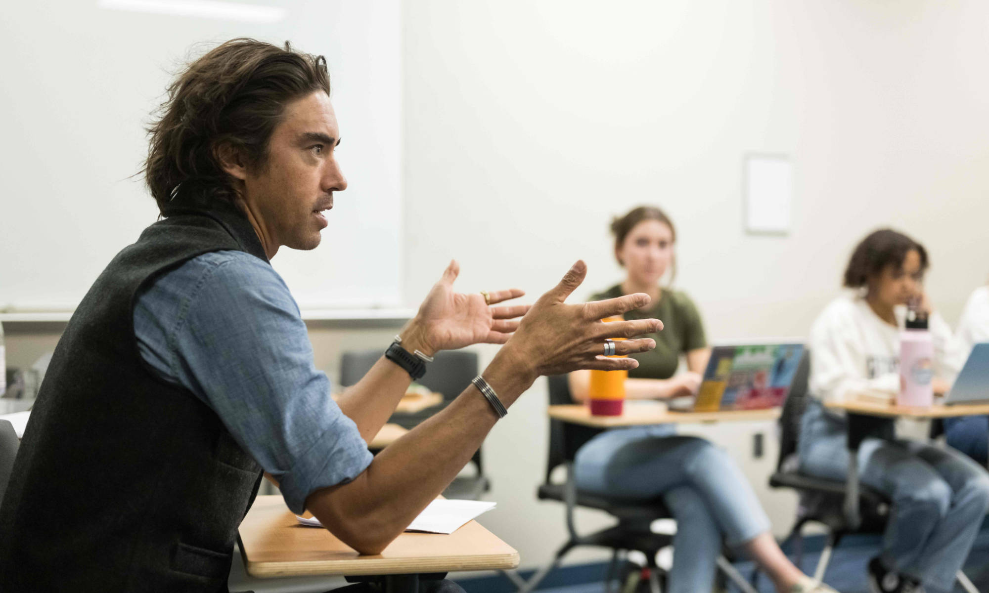 Feature image Jack Downey seated at a desk in a classroom teaching with two students seated with their laptops out of focus in the background.