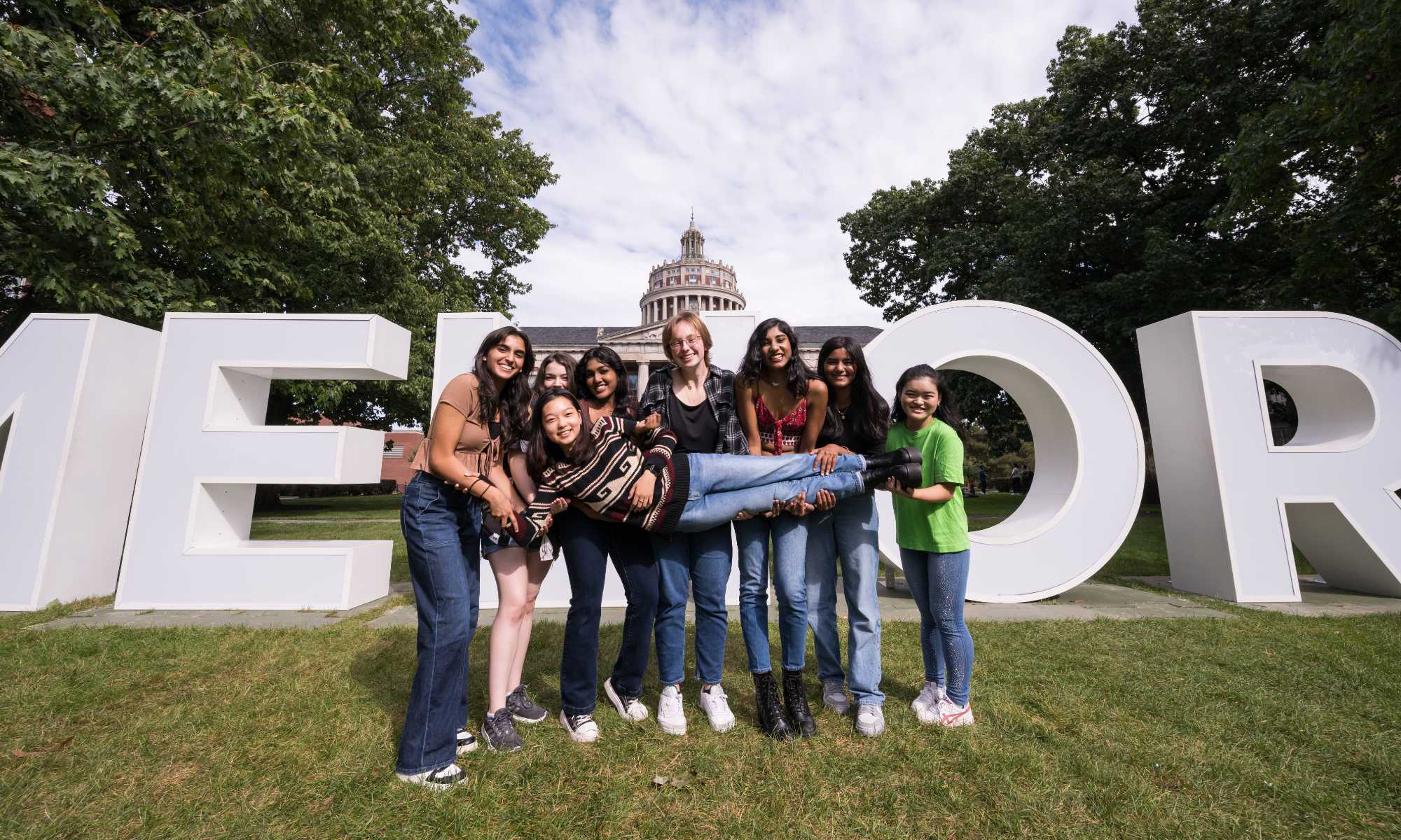 Feature image Six University of Rochester students hold up a seventh in front of the giant letters on the quad during Meliora Weekend 2023.
