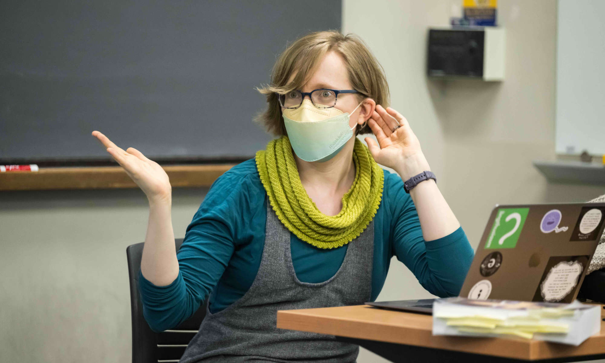 Feature image Whitney Gegg-Harrison wears a mask and gesticulates while seated in front of a laptop at the head of a classroom.