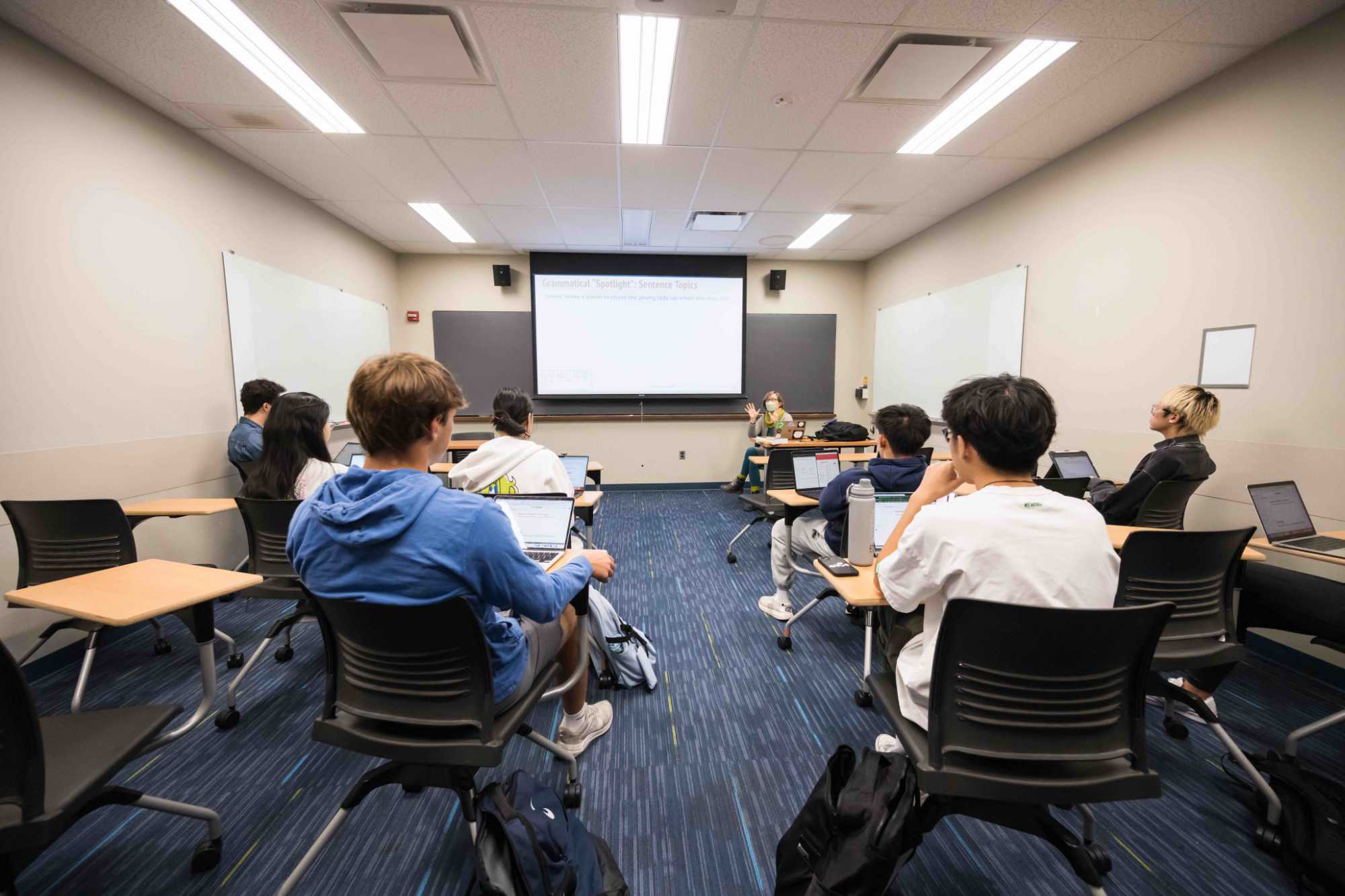 Classroom with about ten students seen from behind looking at the projector and instructor at the front of the room.