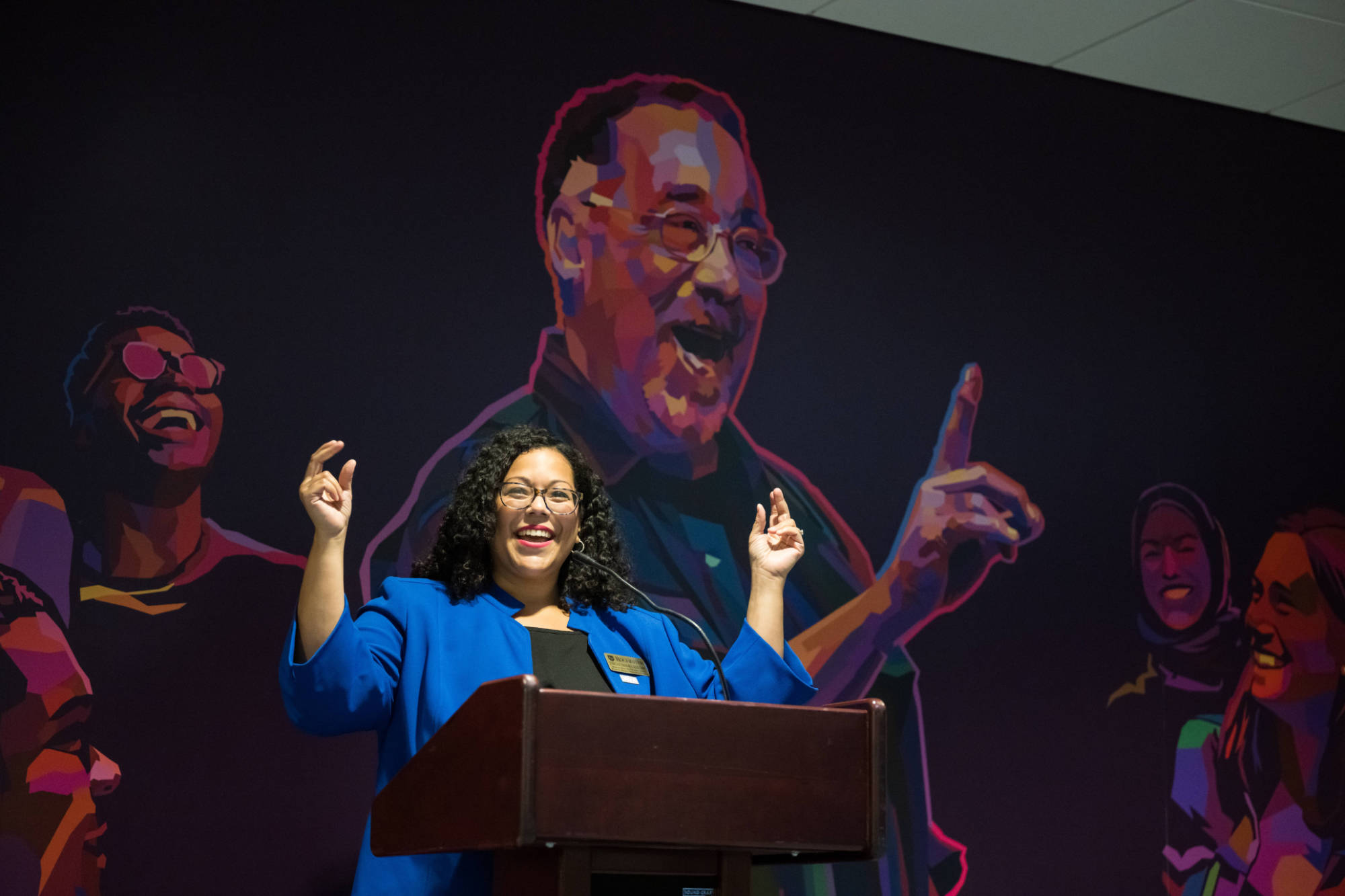 Jessica Guzmán-Rea in front of a podium with both arms raised and a mural of Paul Burgett behind her.