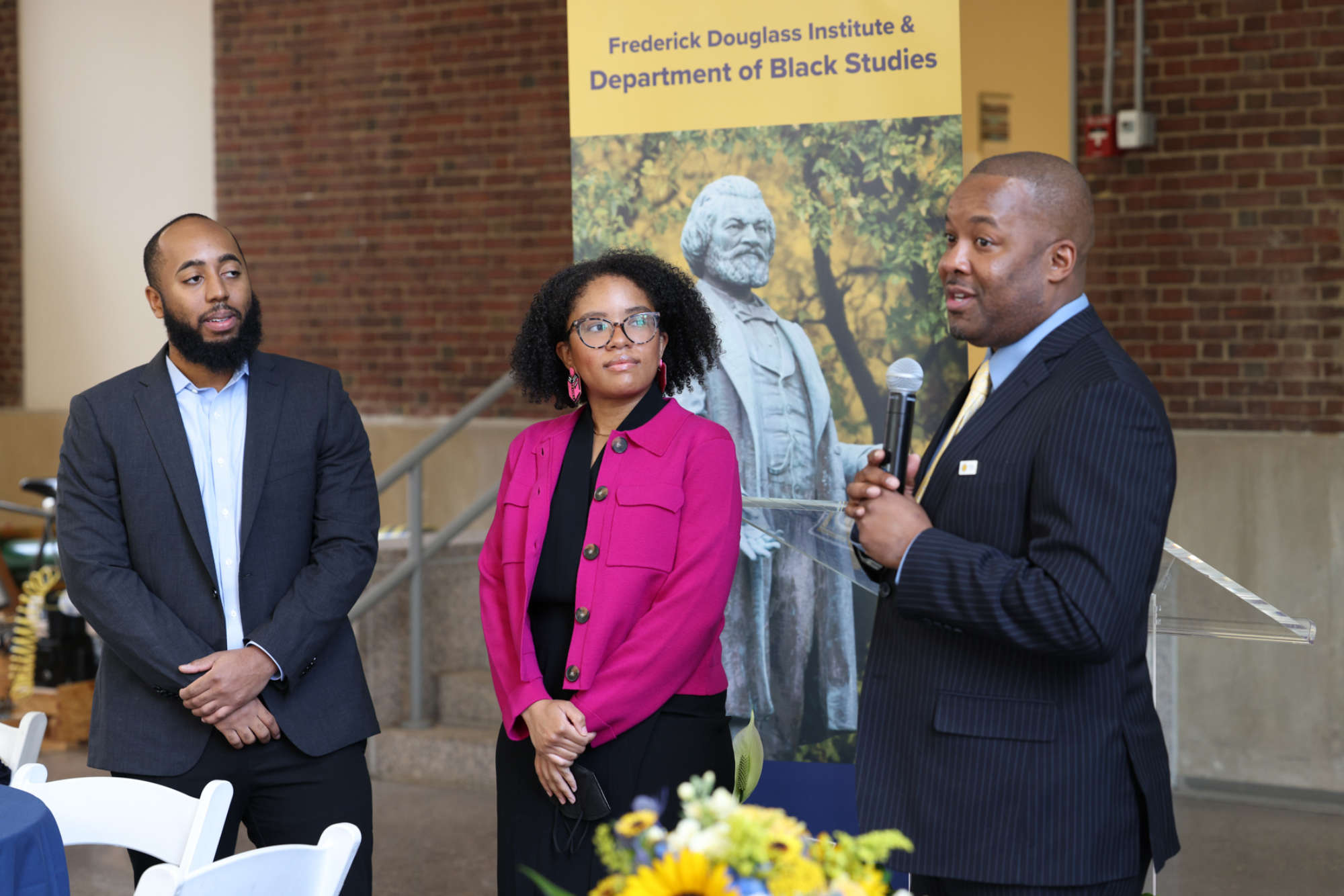 Philip V. McHarris, Jordan Ealey, and Jeffrey McCune Jr. stand in front of signage that says "Department of Black Studies and Frederick Douglass Institute" and shows a statue of Frederick Douglass.