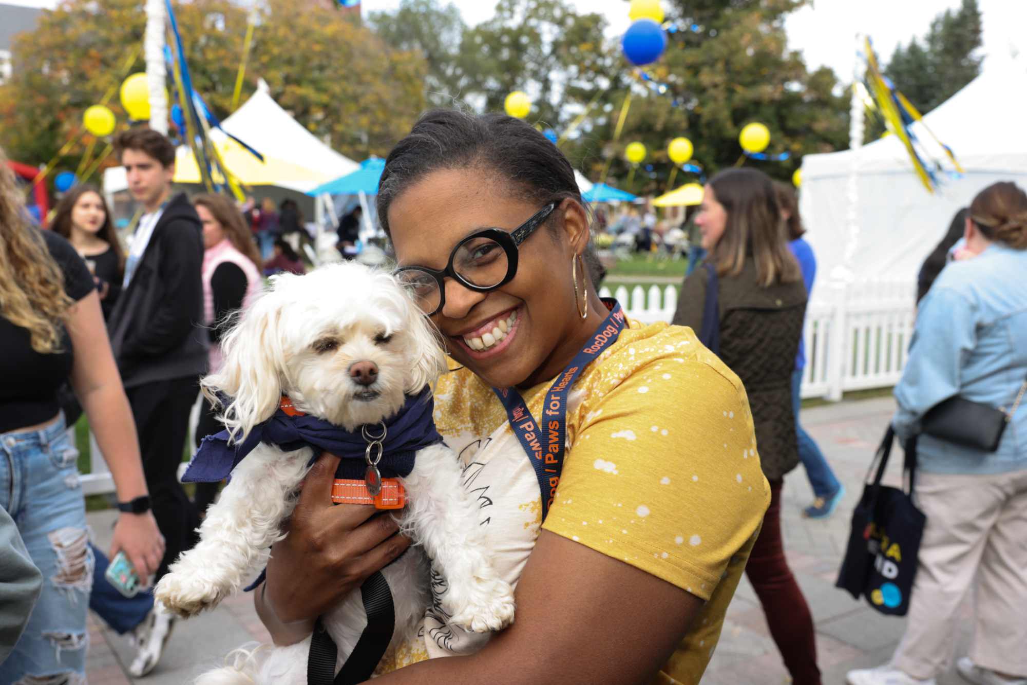 Woman smiles and holds a small dog in Meliora Village with guests around.