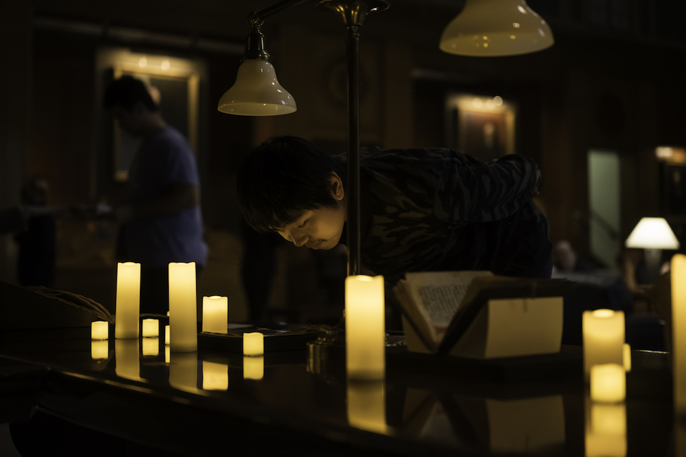 A person leaning over a table with an old manuscript surrounded by electric candles