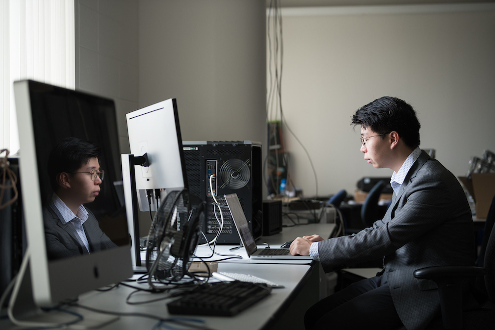 A student in a grey sweater and white button down shirt working at a desk with three computers