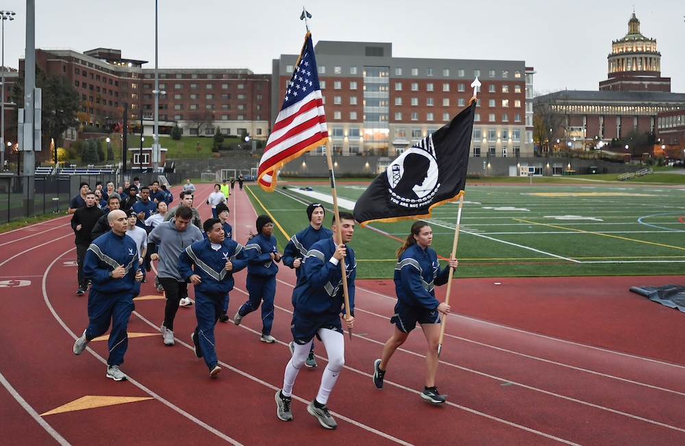 A group of runners in rounding a track. The two runners in the lead carry and US flag and a POW/MIA flag.