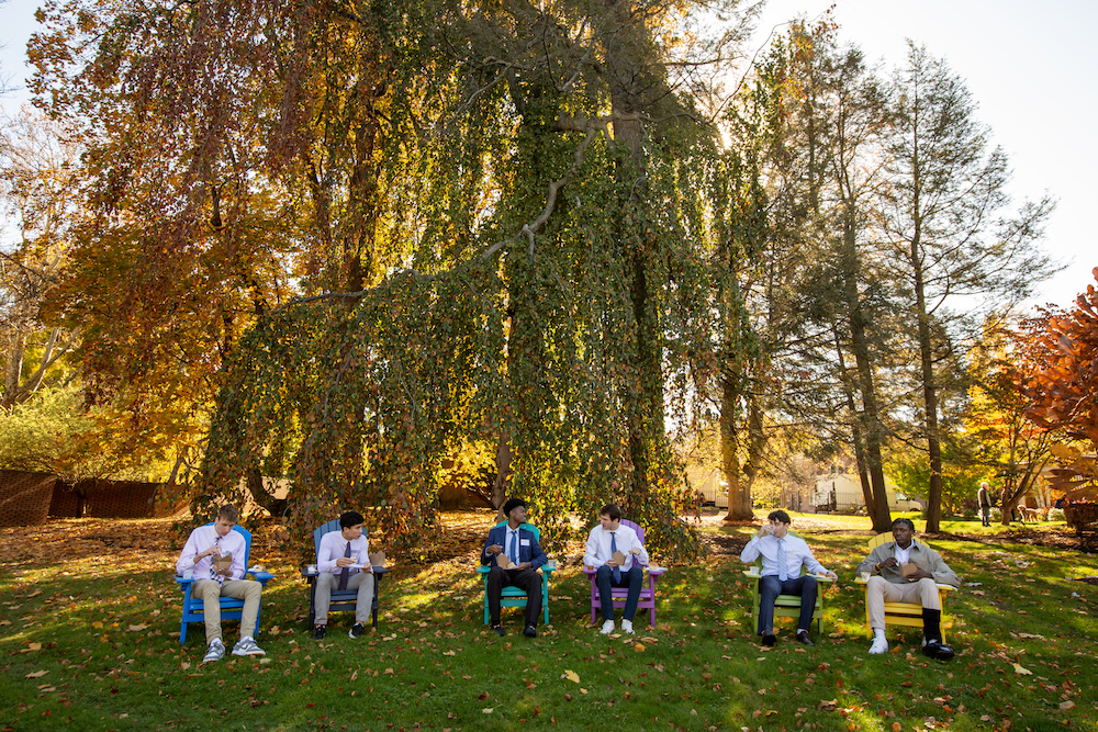Six college-aged men sit on Adirondack chairs, outside on a lawn surrounded by fall foliage