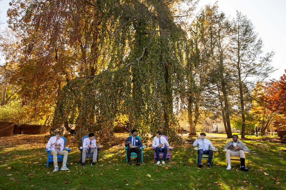 Six college-aged men sit on Adirondack chairs, outside on a lawn surrounded by fall foliage