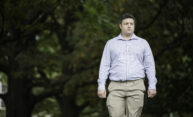 man in a blue button down shirt and beige pants looks off into the distance as he walks on campus.