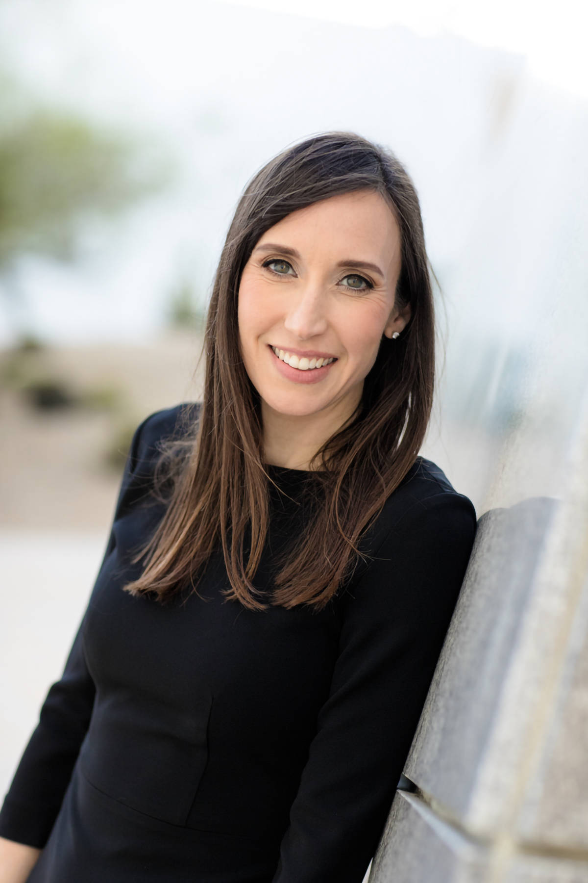 Vertical portrait of Sarah Jesse in black smiling and looking at the camera while leaning against a wall. 