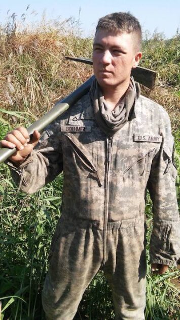 Garrett Boehmer wearing army fatigues holds a large tool over his shoulder.