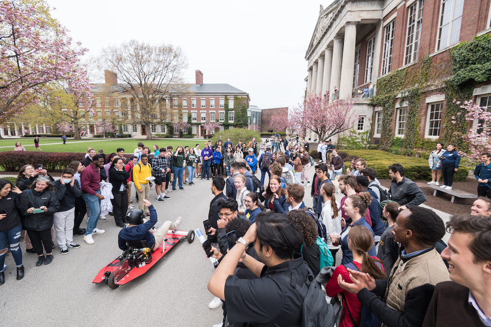 College student in a car powered by a hand drill is cheered on while he races by a group of supporters