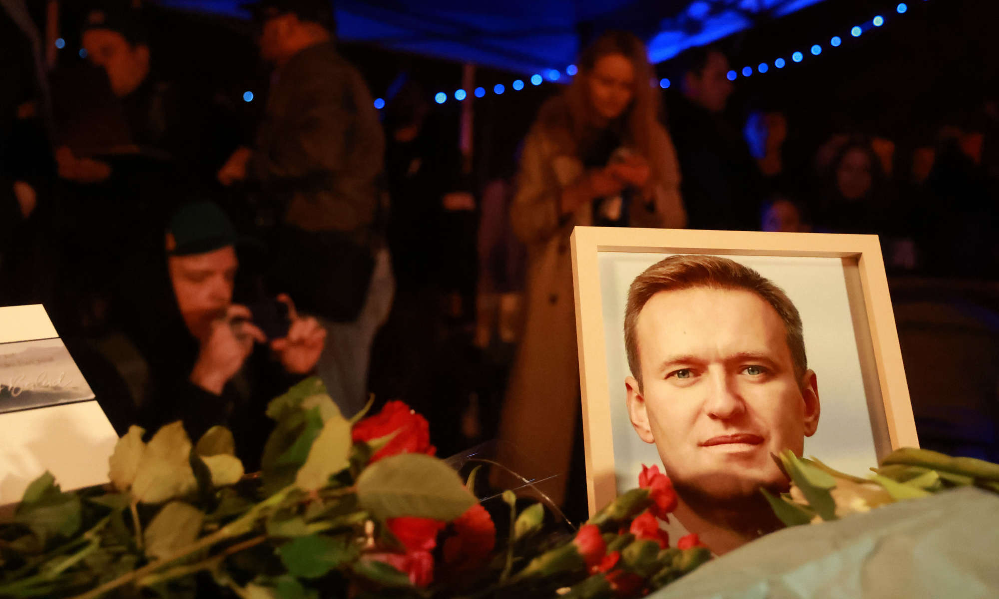 Close up of a framed photo of Alexei Navalny, who led one of the recent opposition movements against Vladimir Putin, surrounded by flowers during a nighttime vigil after reports of his death.