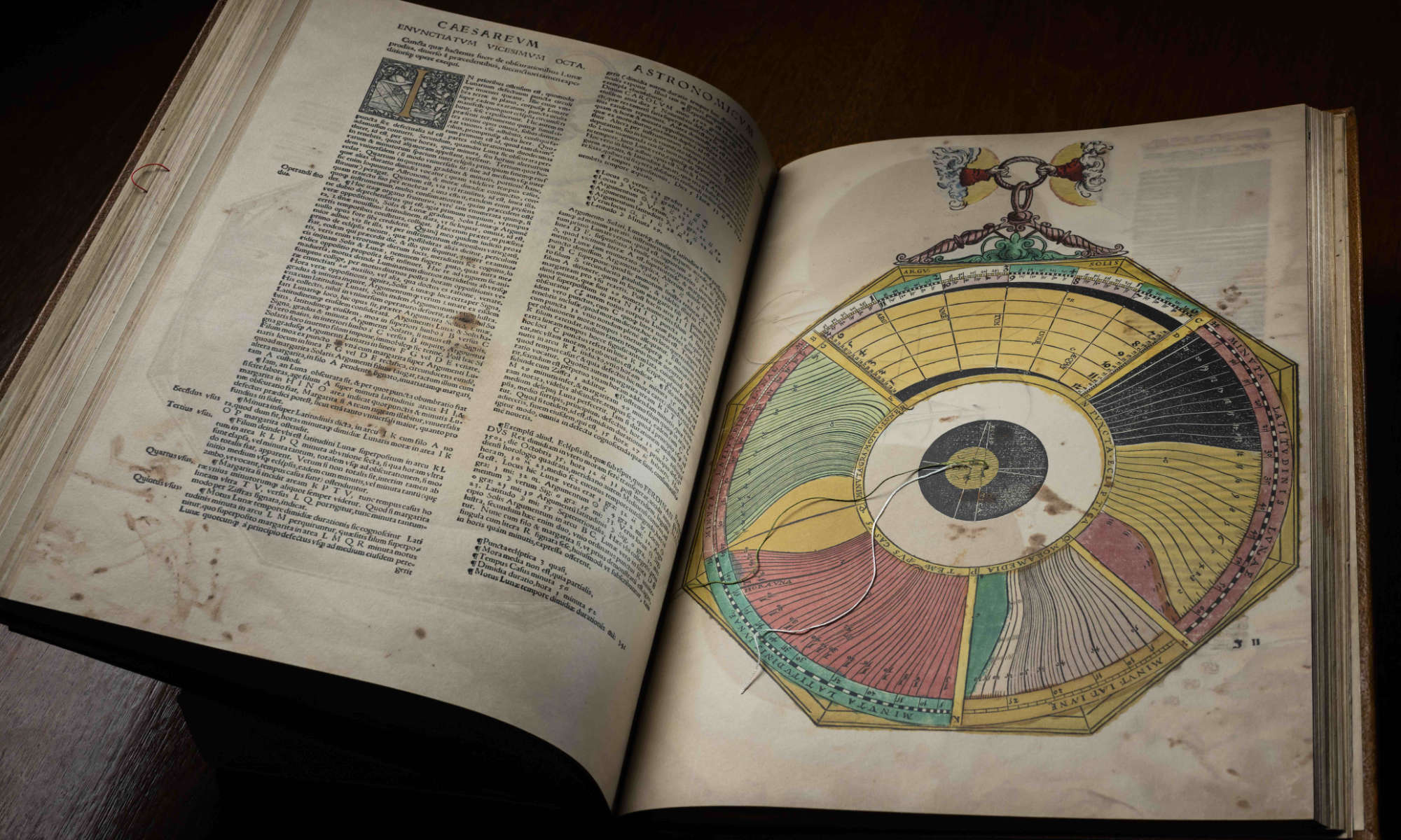 A facsimile of “Astronomicum Caesaream” by Petrus Apianus opened to a spread that shows lots of text on the left page and a colorful compass-like illustration on the right.