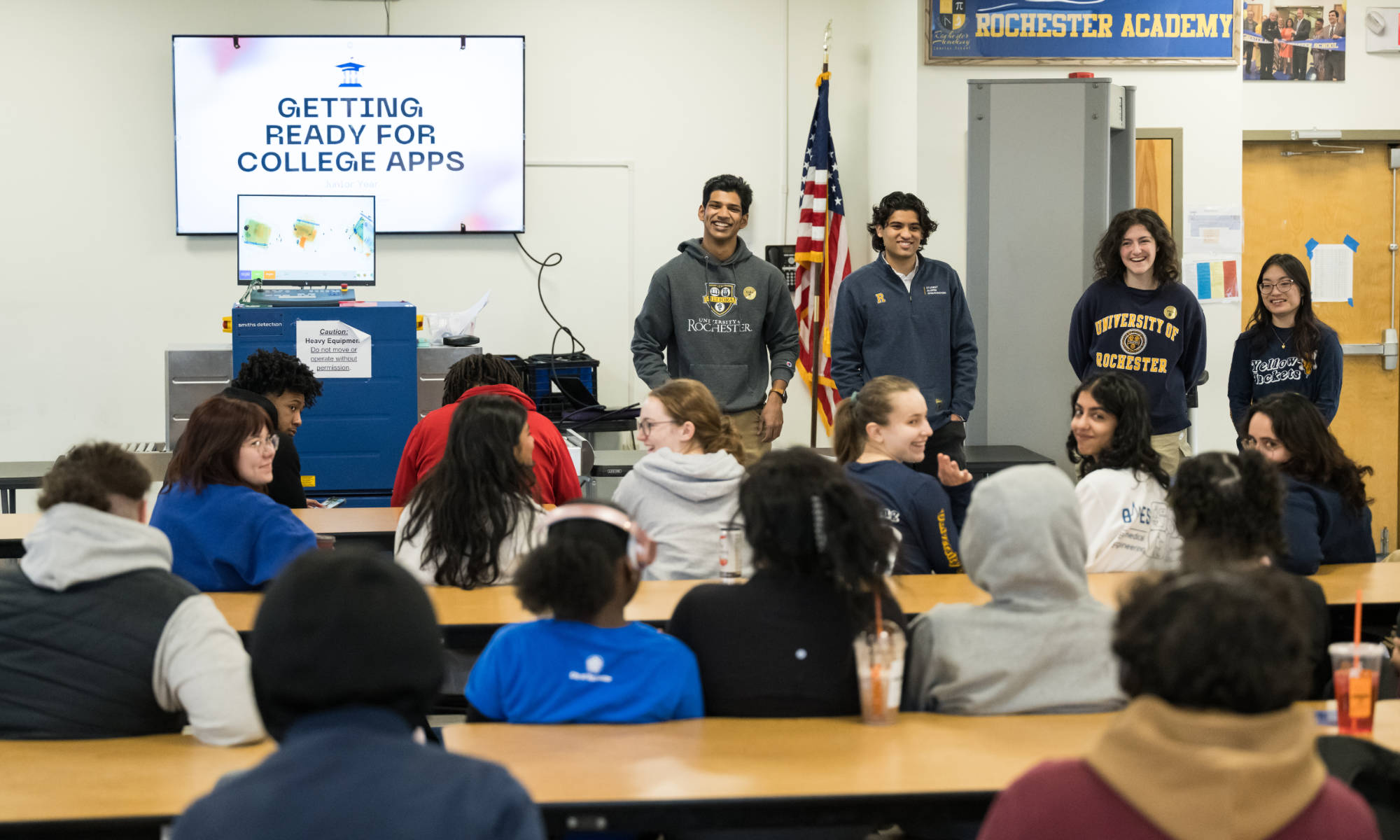 Student educational consultants from Project Level the Field wear University of Rochester gear and stand in front of a room full of high school students with a screen near them that reads "getting ready for college apps."