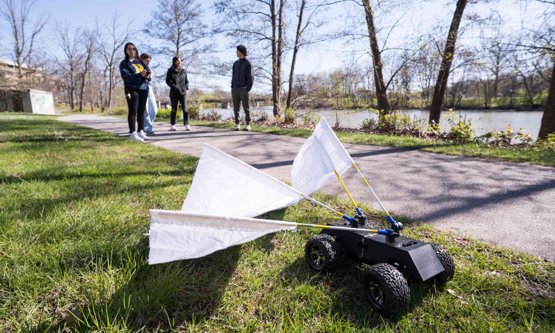 A radio controlled car with white flannel flags attached and four students in the background controlling it on a bike path.