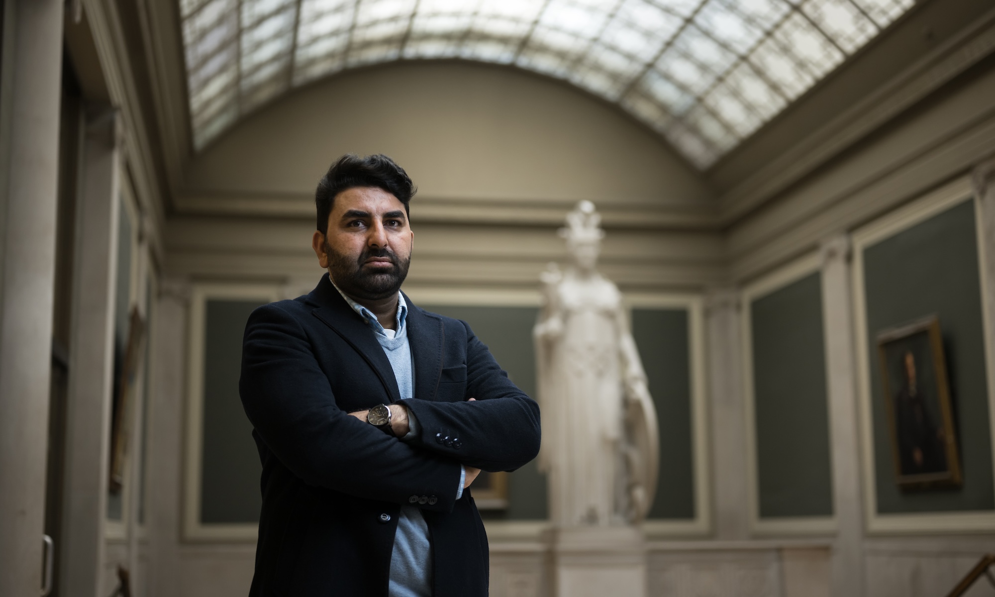 Hameedi standing with arms folded in front of statue in Rush Rhees Library.