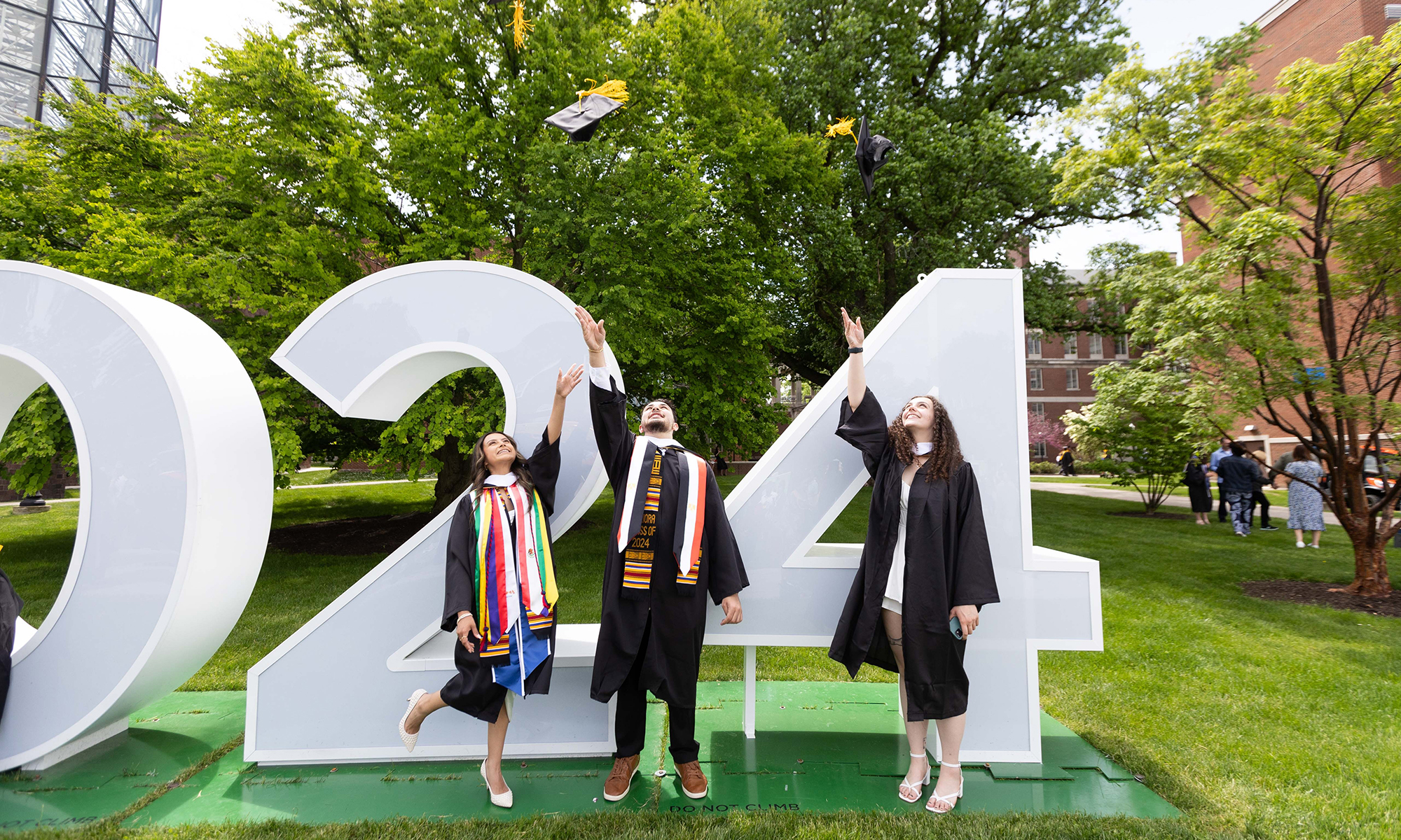 Three students throw caps in the air, standing in front of numbers 24 on quad.