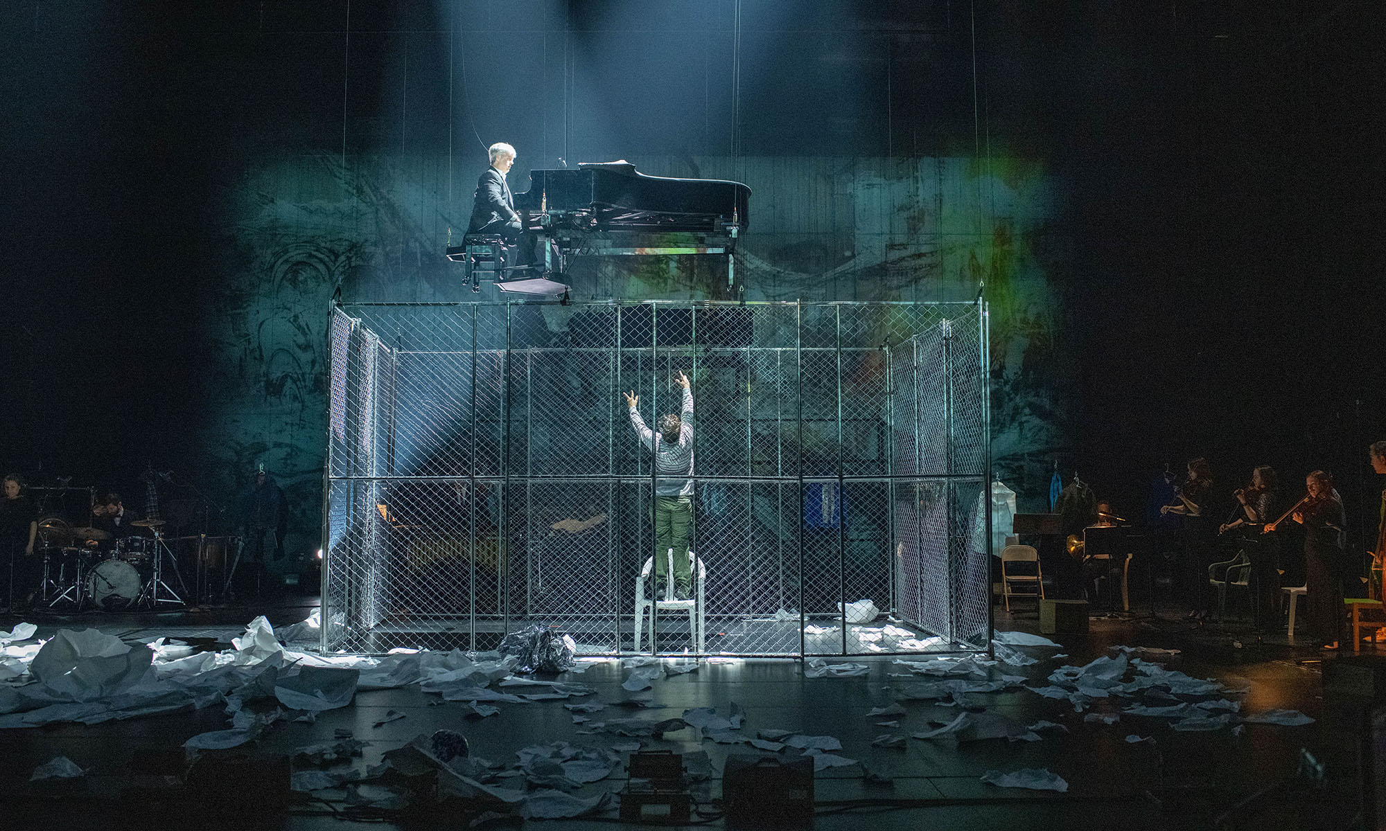 Man in cage reaches up toward piano player in scene from Paper Pianos.