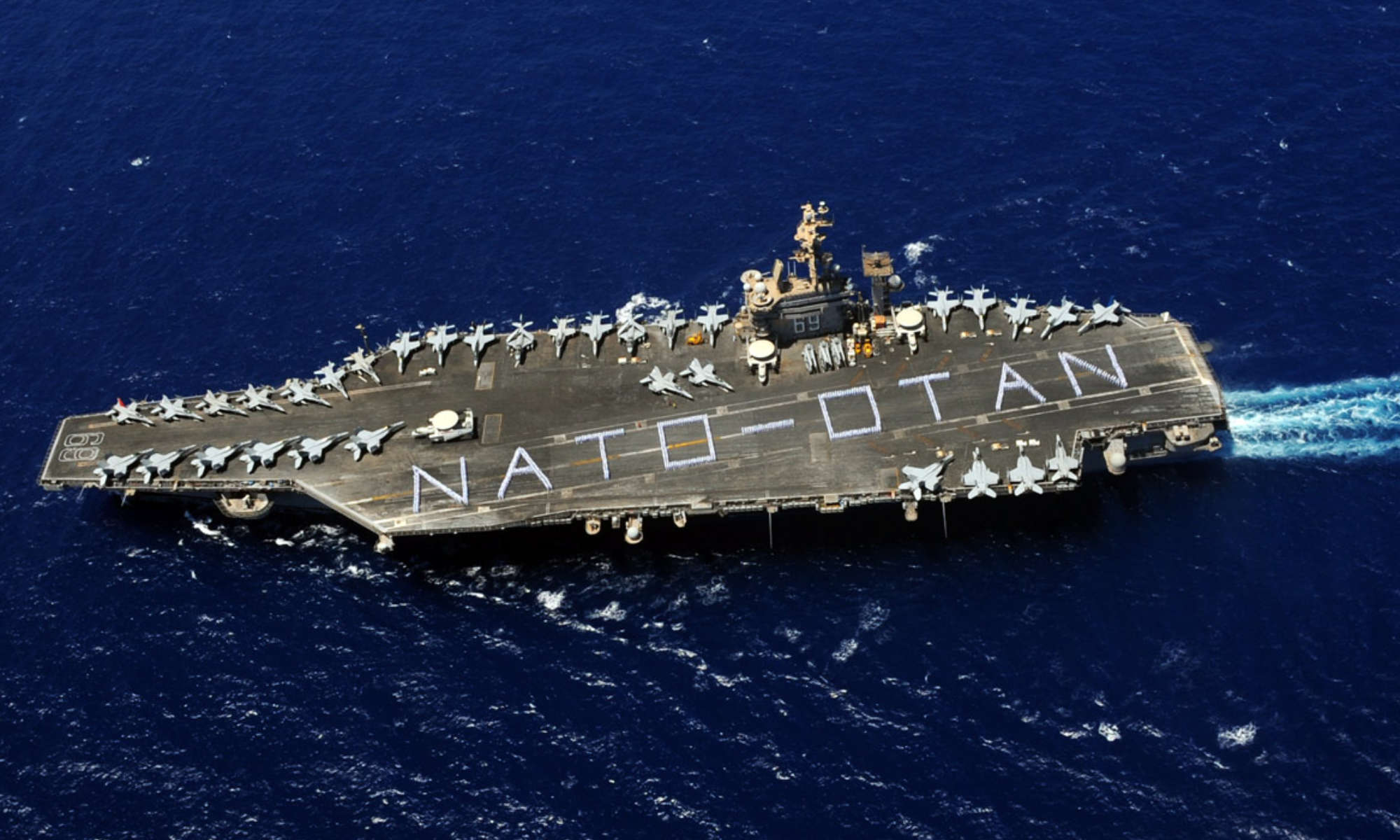 Aerial view of an aircraft carrier at sea with sailors spelling out the words NATO and OTAN.