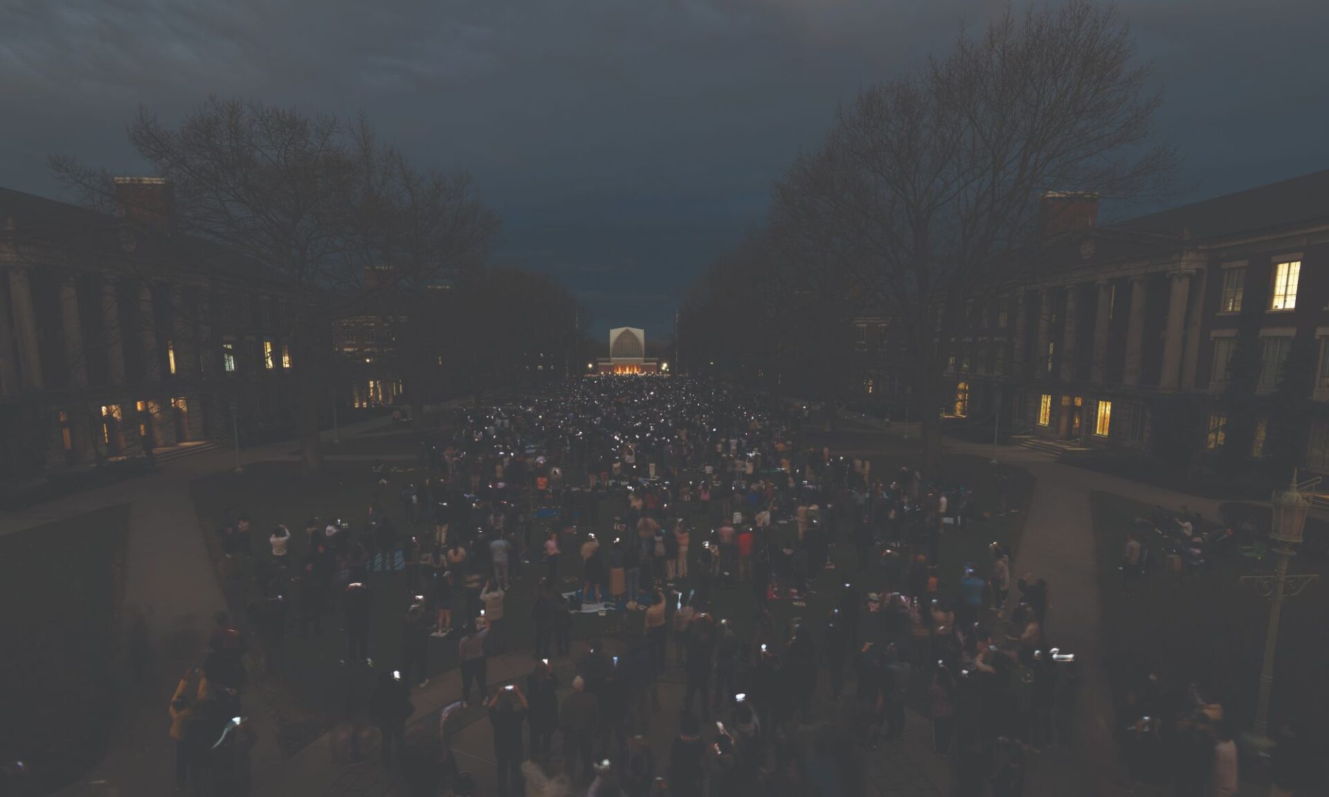 A large crowd of people gathers in an outdoor courtyard at dusk, holding up lit candles or lights. Trees surround the area and buildings with lit windows line both sides. The sky is darkening, and the scene appears peaceful and solemn.