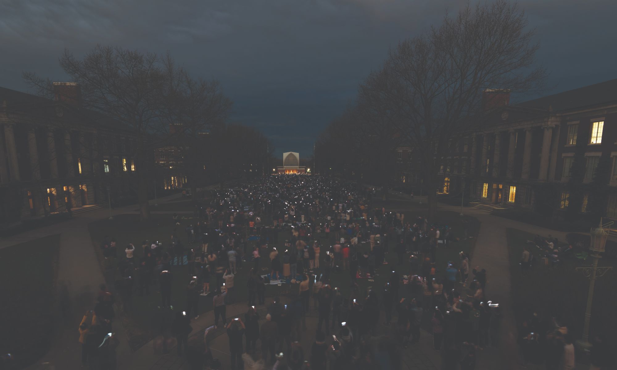 A large crowd of people gathers in an outdoor courtyard at dusk, holding up lit candles or lights. Trees surround the area and buildings with lit windows line both sides. The sky is darkening, and the scene appears peaceful and solemn.