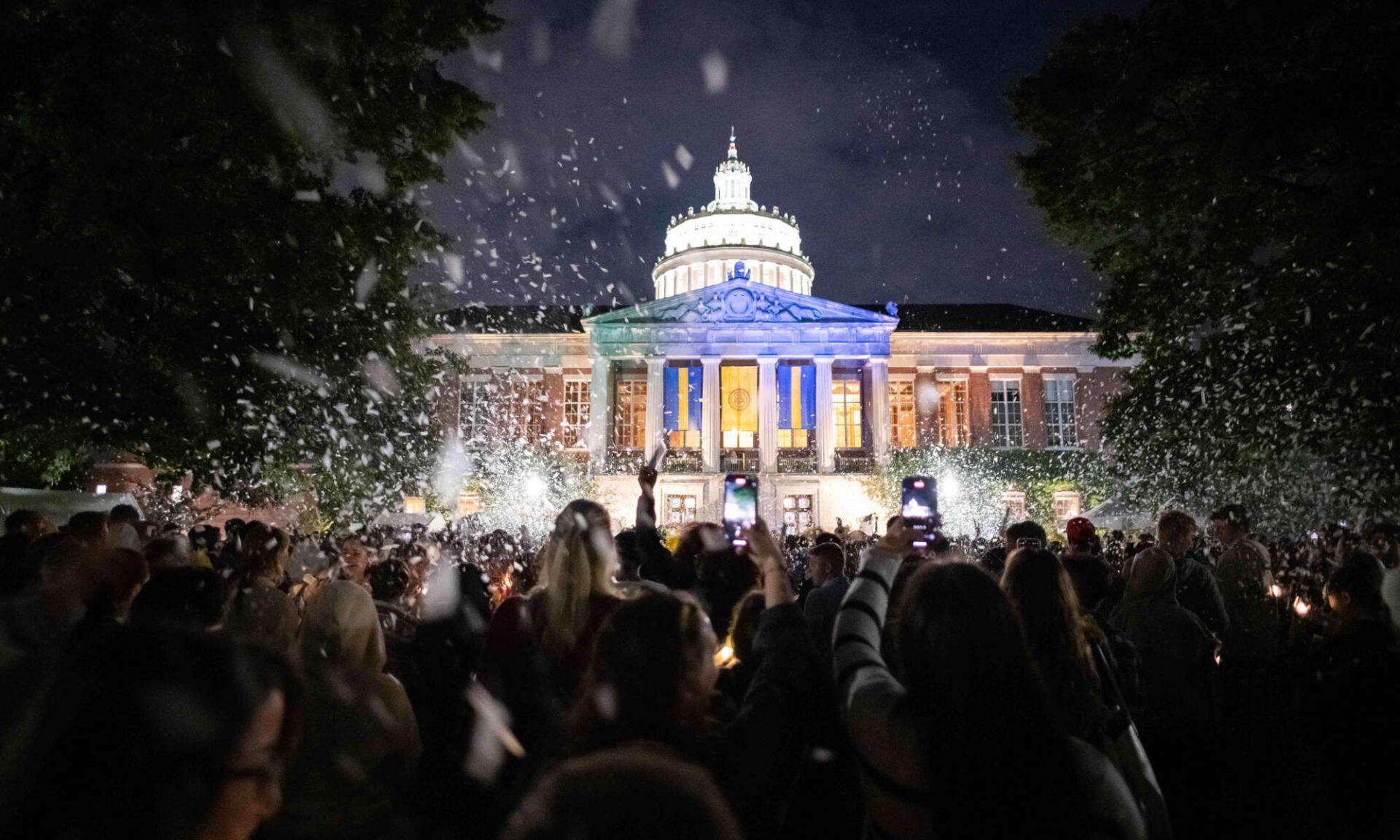 University of Rochester students celebrate, cheer, and throw confetti into the night sky with Rush Rhees Library lit up in the background.