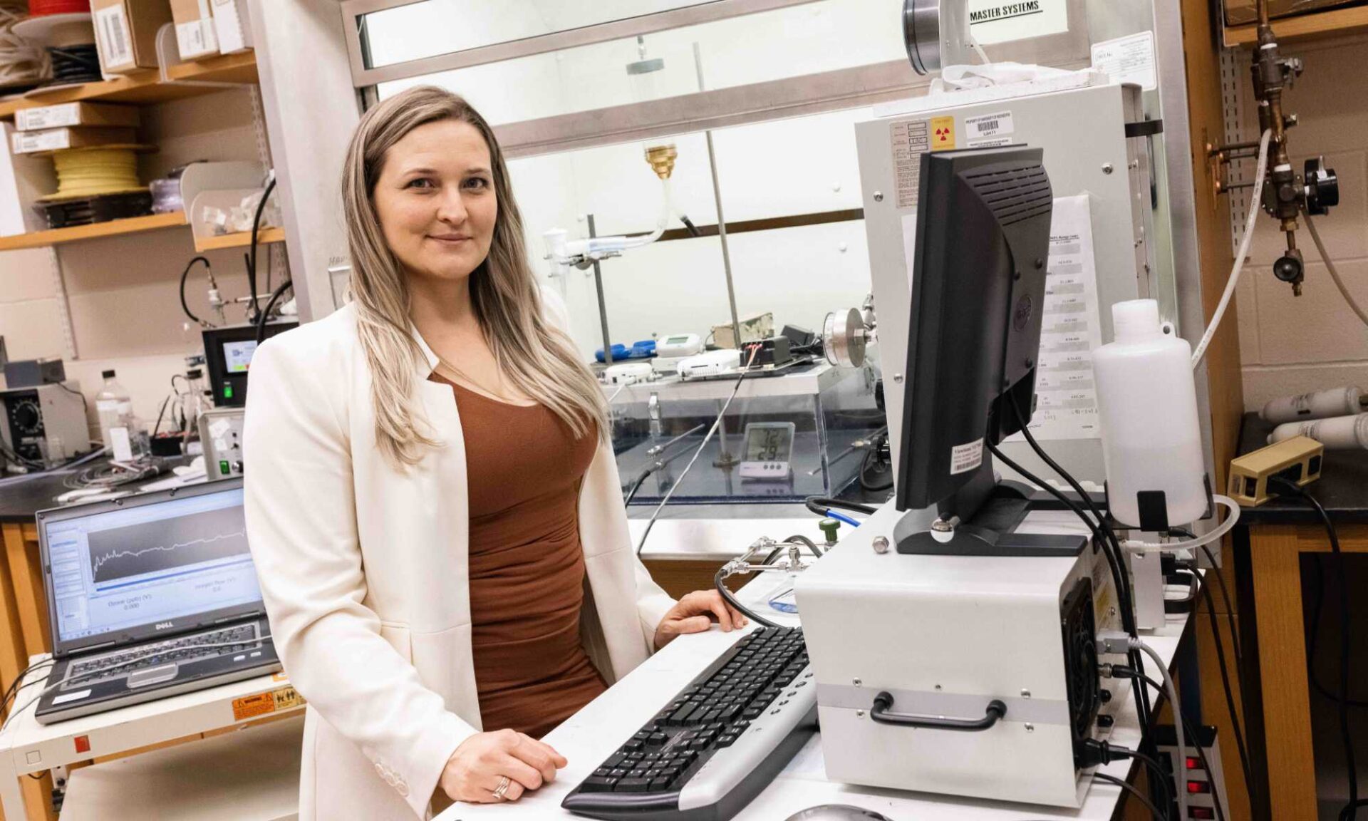 Marissa Sobolewski in a white lab coat stands at a computer station while looking at the camera and smiling.