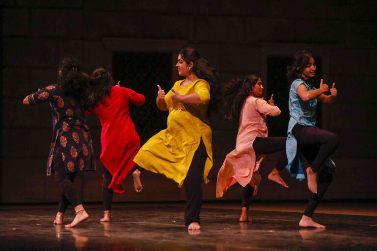 South Asian dancers in brightly colored outfits dance on stage.