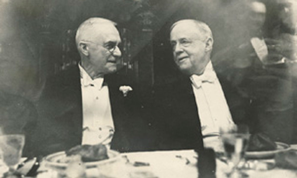 Two elderly men in formal attire seated at a dinner table, engaged in conversation.
