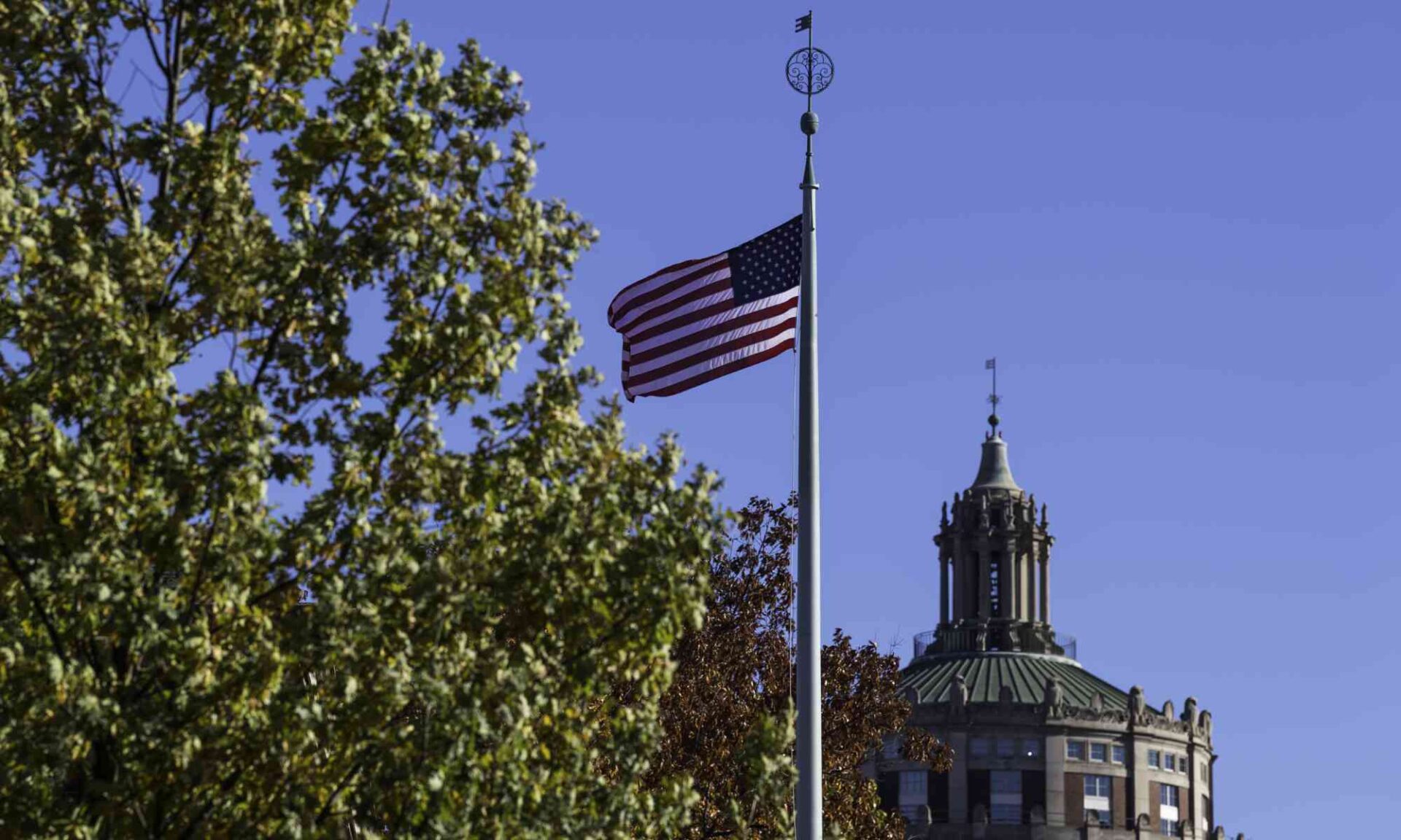 American flag flying with green trees, blue sky, and Rush Rhees Library in the background.