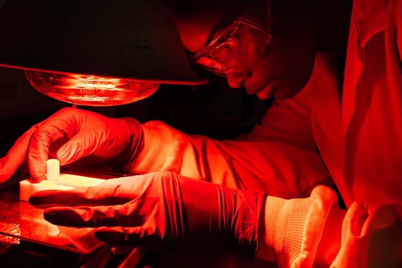 student in protective goggles and gloves places a sample under a light that bathes the whole scene in a red glow.