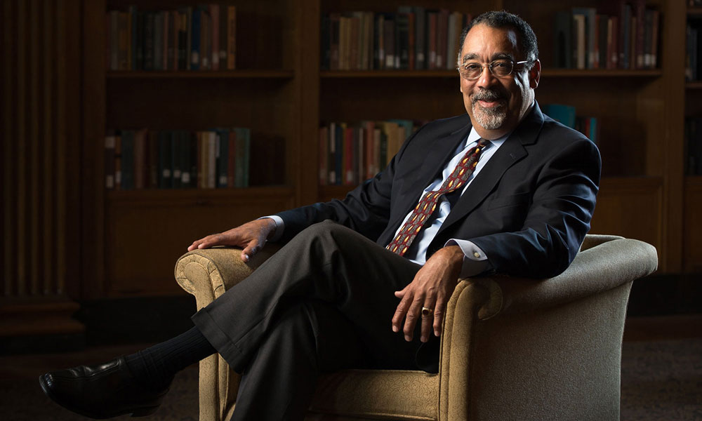 Person in a dark suit seated in a library armchair, with bookshelves in the background.
