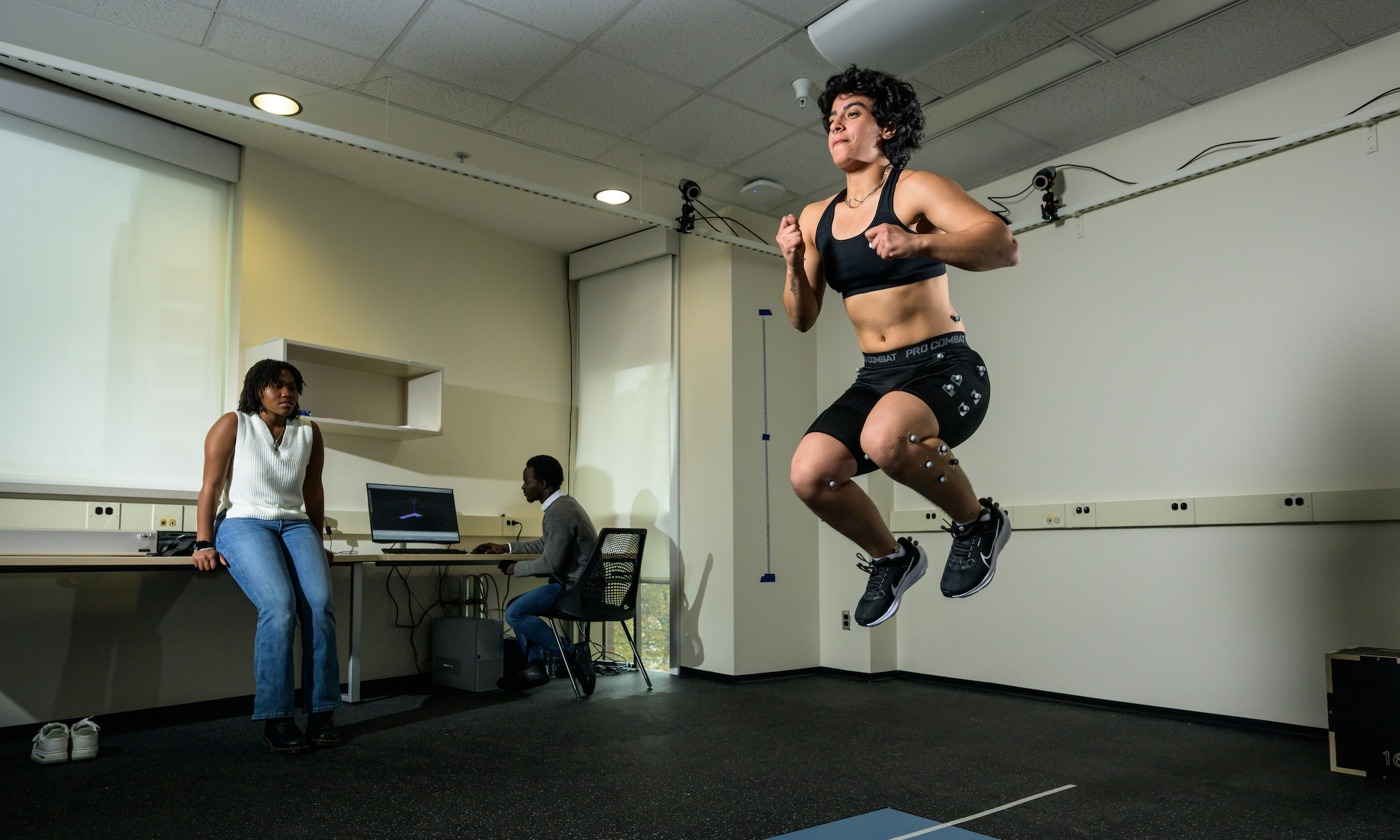 Student performs various movement and balance tasks on a force plate walkway while wearing shoes instrumented with pressure insoles and a suit outfitted with reflective markers for use in collection of biomechanics data