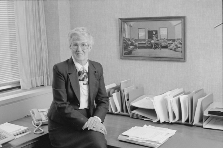 Loretta Ford, former School of Nursing dean, is depicted in a suit and tie, seated at a desk in a professional setting.